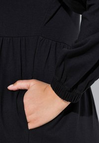 Czarna sukienka z marszczonym paskiem w talii i długimi rękawami. Posiada boczną kieszeń i elastyczne mankiety. Gładka, miękka tekstura.