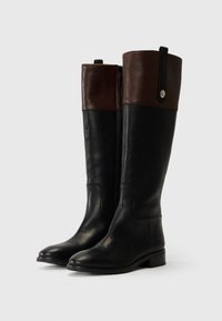 Bottes noires en cuir hautes avec un revers en cuir marron, dotées d'une fermeture éclair sur le côté et d'un accent rond en métal argenté sur le revers. Semelle plate.