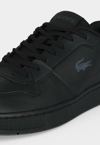 Μαύρο δερμάτινο sneaker Lacoste με μαύρα κορδόνια και το λογότυπο κροκόδειλου της μάρκας στο πλάι και κείμενο "Lacoste" στη γλώσσα.