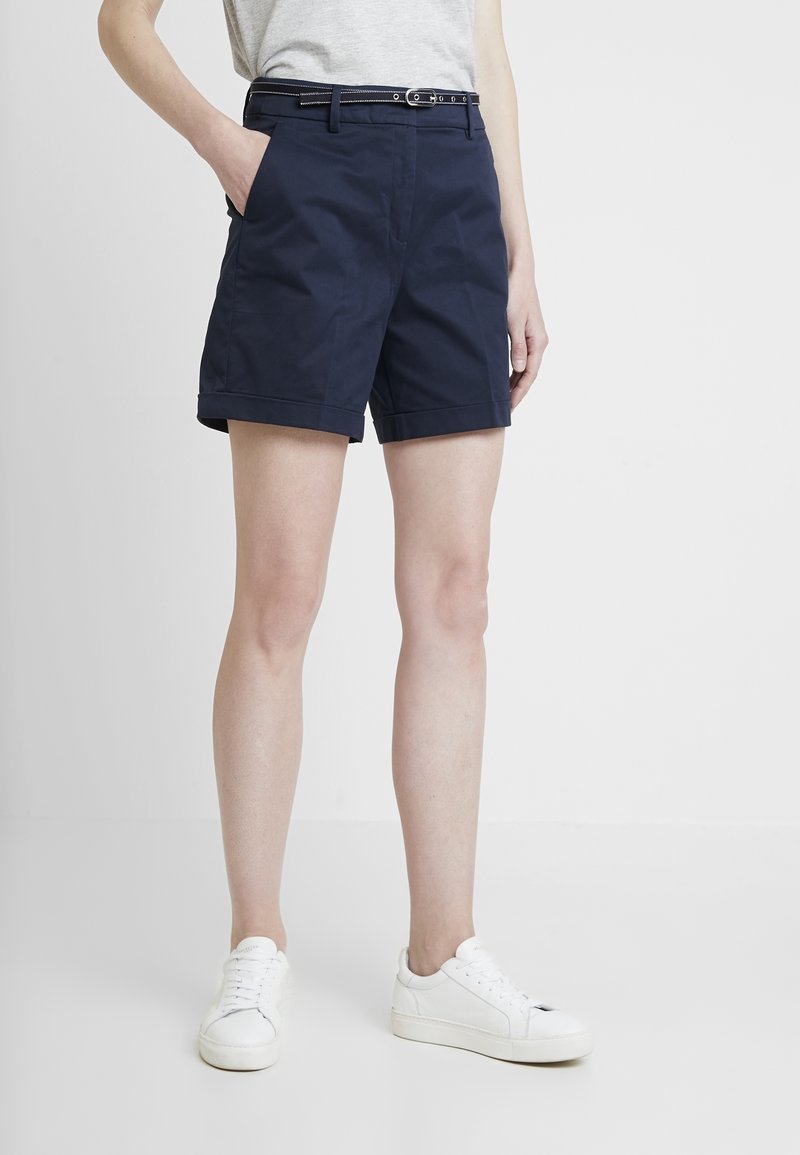 Personne portant un short bleu marine à ourlet retroussé avec une ceinture noire, des baskets blanches et un haut sans manches gris clair, debout devant un fond blanc.