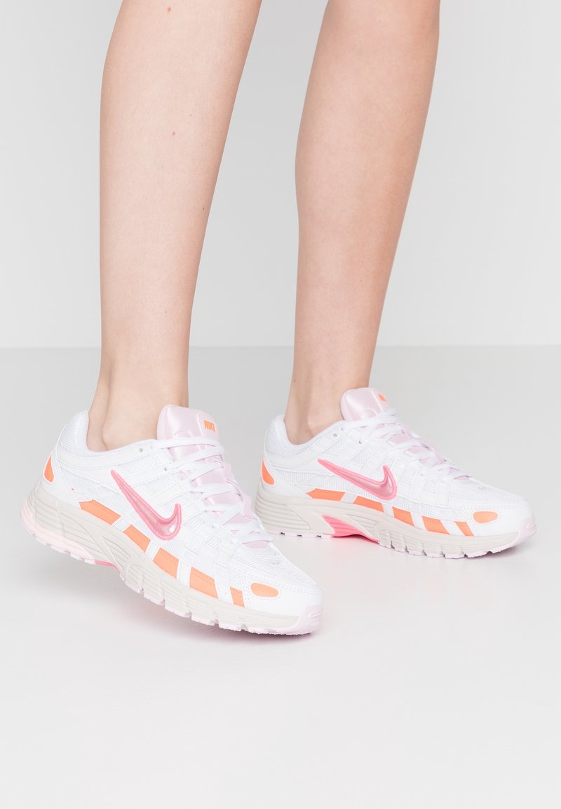 Personne portant des chaussures de sport blanches avec des accents roses et orange, debout sur un sol gris clair devant un fond uni.