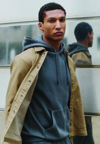 Veste marron portée sur un sweat à capuche gris, doté d'une poche kangourou et de cordons de serrage. Le modèle pose devant une surface réfléchissante.