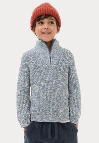 Maglione lavorato a maglia blu chiaro con colletto alto e mezza zip; motivo testurizzato; indossato sopra una camicia bianca; abbinato a pantaloni in velluto a coste blu scuro e un berretto rosso.