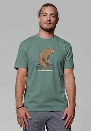 Grünes Baumwoll-T-Shirt mit einer detaillierten Illustration eines Bären, der ein rotes Fahrrad fährt. Kurze Ärmel und lockere Passform.