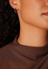 Gouden ringoorbel, kleine maat, met een gepolijste afwerking. Draagt dicht bij het oorlel, gecombineerd met een bruin stoffen top die een gladde textuur laat zien.