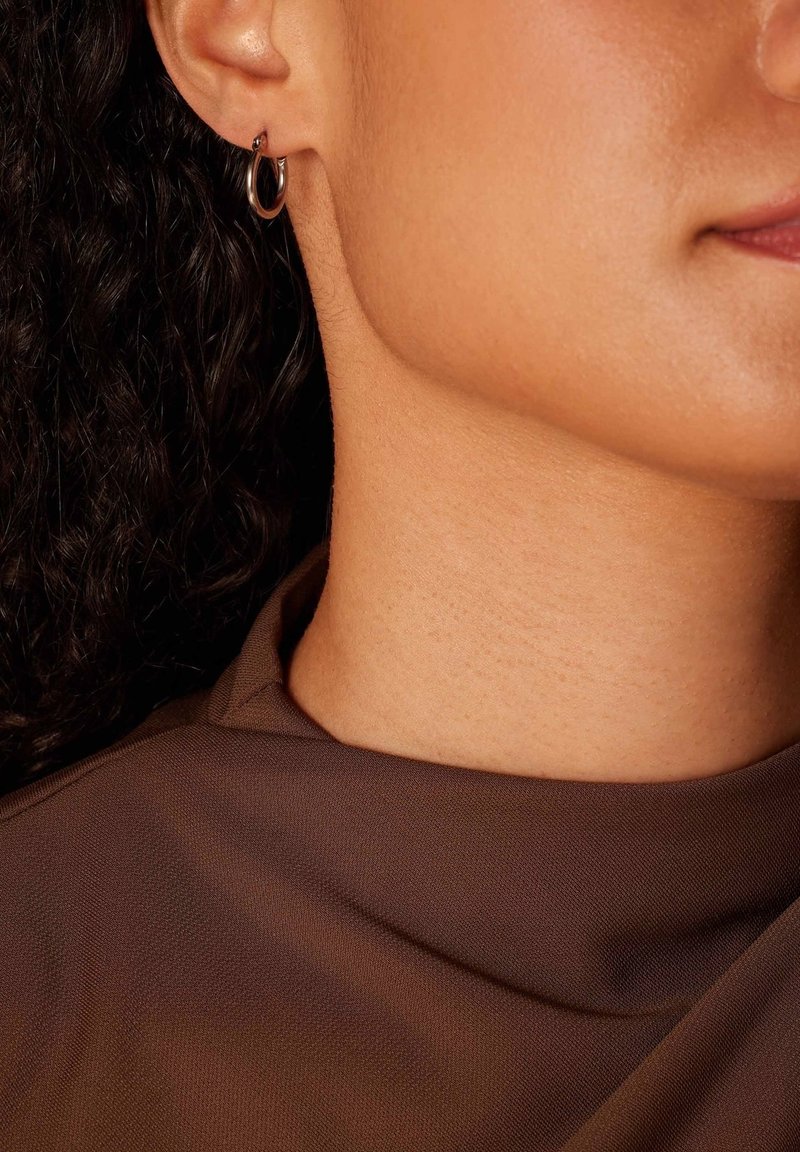 Gouden ringoorbel, kleine maat, met een gepolijste afwerking. Draagt dicht bij het oorlel, gecombineerd met een bruin stoffen top die een gladde textuur laat zien.