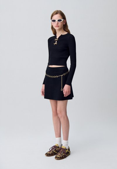 Top de punto negro de manga larga con acentos de lazo dorado, combinado con una minifalda negra que presenta un cinturón de cadena dorada. Zapatillas impresas completan el look.