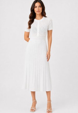 Vrouw staand, draagt een wit kort mouwtjes knoopjes top met kantpatroon en een witte geplooide midi rok met sandalen met bandjes.