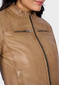 Veste en cuir brun avec un col haut, dotée de deux poches zippées et d'une texture lisse. Le design est ajusté avec des détails de surpiqûres subtils.