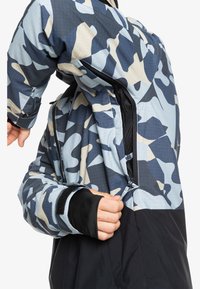 Farverig camouflagejakke med en sort nederste sektion, der har en lynlås, meshforing og tommelhuller på ærmerne. Vandafvisende materiale.