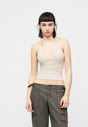 Jeune femme aux cheveux courts et foncés, portant un haut beige à col licou et un pantalon cargo gris ample, sur un fond clair uni.