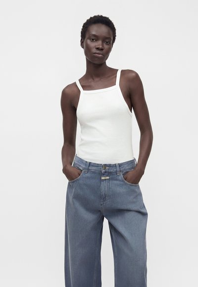 Femme aux cheveux courts portant un haut blanc sans manches et un jean bleu ample, debout avec les mains dans les poches devant un fond neutre.