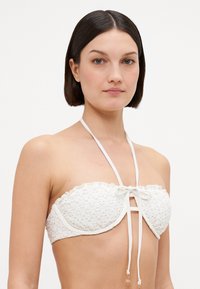 Bela strapless bikini zgornji del iz teksturirane tkanine, okrašen s čipkastim robom in sprednjo mašno, z tankimi nastavljivimi trakovi okoli vratu.