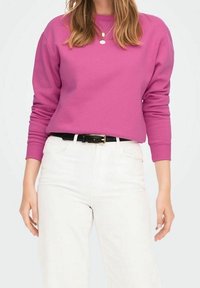 Sudadera fucsia de manga larga, escote redondo y puños acanalados. Combinada con vaqueros blancos y cinturón negro con hebilla metálica.