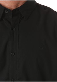 Acercamiento de una persona que lleva una camisa negra de botones con cuello y un sutil logo bordado en el lado izquierdo del pecho.