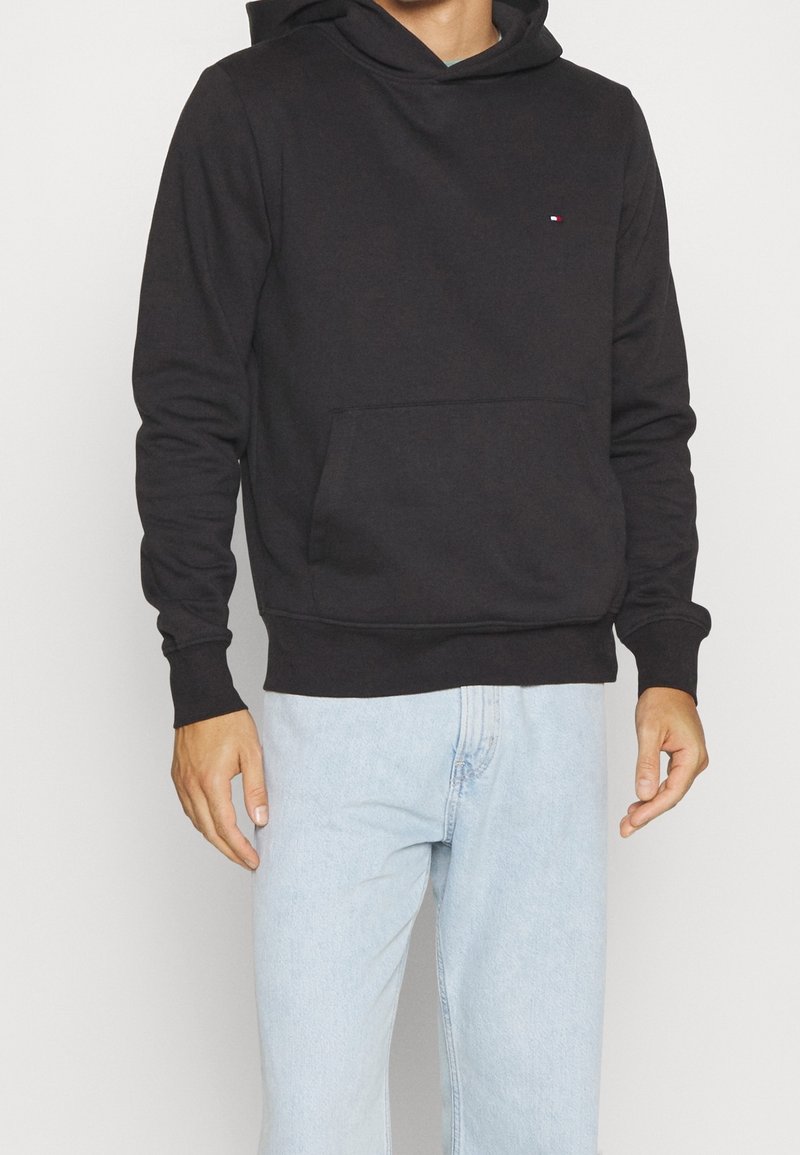 Homme portant un sweat à capuche noir uni avec poche avant et un jean bleu clair, debout devant un fond blanc.