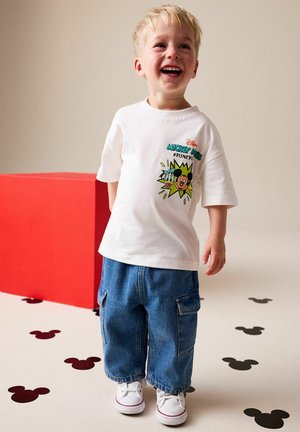 Smilende lillebarn iført hvid t-shirt med Mickey Mouse-grafik, brede blå cargobukser og hvide sneakers, stående på gulv med sorte og røde Mickey Mouse-figurer.