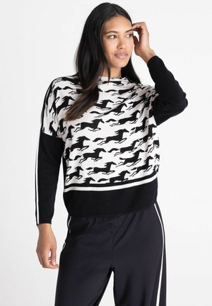 Vrouw draagt een zwart-witte trui met een patroon van een rennend paard, zwarte broek met witte strepen, poseert met één hand nabij haar haar.