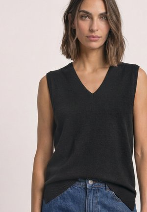 Donna con capelli castani fino alle spalle, che indossa un maglione nero senza maniche con scollo a V e jeans blu, rivolta frontalmente su uno sfondo semplice.