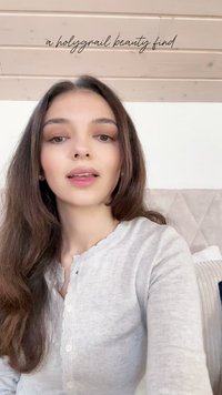 Helle graue Strickjacke mit einem gewellten Kragen, getragen von einer Person mit langen, dunklen Haaren. Sanfter, neutraler Hintergrund mit strukturierten Kissen.