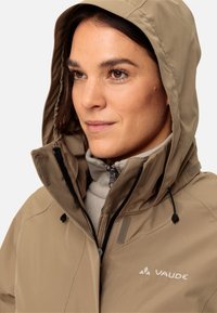 Giacca impermeabile beige con cappuccio, dotata di un colletto alto, cerniera frontale e cordini regolabili. Logo del marchio visibile sul petto.