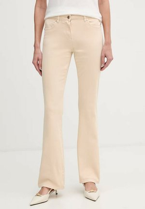 Donna che indossa pantaloni beige a vita alta, leggermente svasati con tasche frontali e décolleté beige con punta appuntita e dettagli dorati.