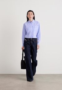 Camicia azzurra chiaro a maniche lunghe con due tasche sul petto, abbinata a jeans a gamba larga blu scuro. Una borsa nera è tenuta nella mano sinistra.