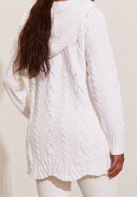 Osoba z długimi brązowymi włosami, ubrana w białą sweter z kapturem o splocie warkocza i białe spodnie, widziana z tyłu na neutralnym tle.