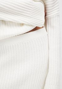 Maglione bianco a coste con una vestibilità comoda. Presenta un tessuto strutturato e polsini larghi, indossato su un fondo in maglia a coste coordinato.