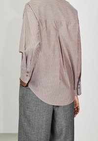 Personne portant une chemise beige ample à manches longues avec des rayures verticales blanches et un pantalon gris à jambes larges, vue de dos.