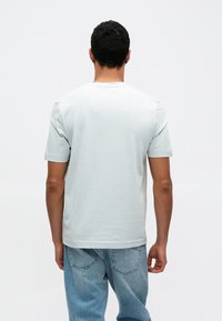 Světlé šedé tričko s krátkými rukávy, kulatým výstřihem, hladkou texturou, nošené s modrými džínami. Pohled zezadu ukazuje uvolněný střih.