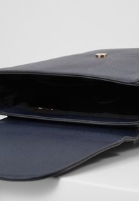 Marinblå läderclutch med en texturerad yta, utrustad med en magnetisk knappstängning och en stilren, minimalistisk design. Inredningen är svart.