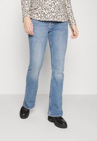 Jeans de ganga azul claro, com corte flared, apresentando um design clássico de cinco bolsos e um desbotado subtil, combinados com calçado preto.