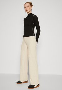Top nero a maniche lunghe con texture a coste e accenti di cucitura bianca, abbinato a pantaloni larghi beige chiaro e scarpe sportive nere.