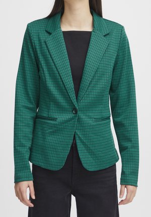 Femme portant un blazer vert à carreaux ajusté avec un seul bouton, associé à un top noir et un pantalon, sur un fond blanc uni.