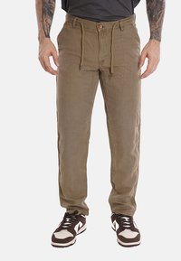 Pantaloni verde oliva da uomo con vita regolabile tramite cordoncino, tasche frontali e un tessuto morbido e strutturato. Abbinati a scarpe da ginnastica marroni e bianche.