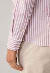 Camicia a maniche lunghe a righe rosa e bianche, con polsini abbottonati e orlo curvo. Il tessuto sembra leggero e ha una texture liscia.