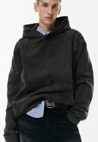 Donkergrijze oversized hoodie met een voorzak, van katoen, met ribgebreide boorden, gelaagd over een lichtblauw gestreept shirt en zwarte spijkerbroek.