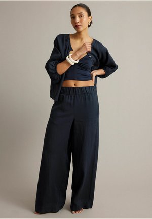 Jonge vrouw staat op blote voeten, draagt losse zwarte broek, een zwarte cropped top, een bijpassend gebreid vest met knopen en witte armbanden om één pols.