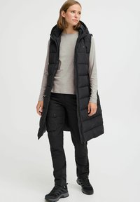 Schwarze gepolsterte Weste mit Kapuze, im langen Design. Kombiniert mit einem grauen Langarmshirt und schwarzen Hosen, getragen mit robusten Schuhen.