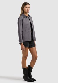 Femeie purtând o jachetă gri cu nasturi, pantaloni scurți negri și cizme negre până la jumătatea gambei, stând în picioare pe un fundal simplu, deschis la culoare.