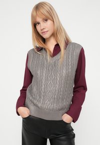 Harmaa neuleliivi, jossa on kaapelikuvio, V-niskamuotoilu, päällä marooni kauluspaita, yhdistettynä mustiin nahkahousuihin.