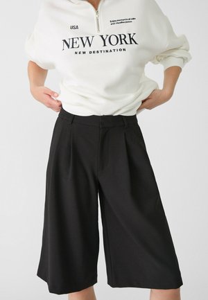 Personne portant un haut blanc à manches longues avec le texte New York et un short noir large arrivant aux genoux, les mains ajustant le short à la taille.