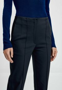 Close-up van een persoon in zwart op maat gemaakte broeken en een langmouw navy-blauw top, met de handen ontspannen aan de zijkanten tegen een effen achtergrond.