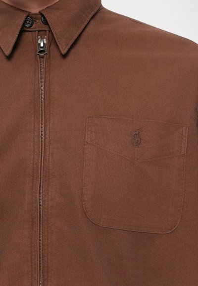 Chemise brune zippée avec col et poche poitrine, ornée d'un logo brodé représentant un joueur de polo à cheval.