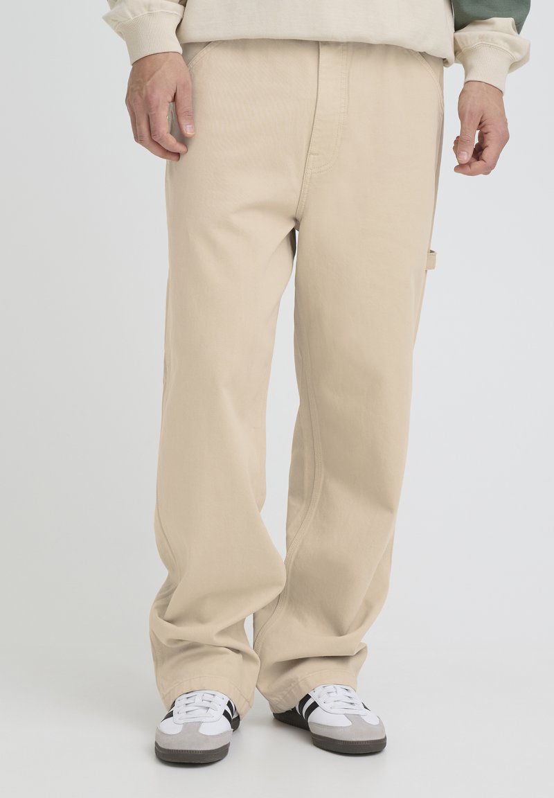 Homme portant un pantalon beige ample et des baskets blanches à rayures noires, debout devant un fond clair uni.