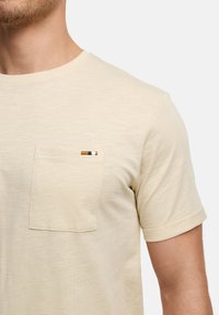 Camiseta de manga corta color beige hecha de un tejido suave, con un bolsillo en el pecho izquierdo que presenta un acento a rayas en azul marino y naranja.