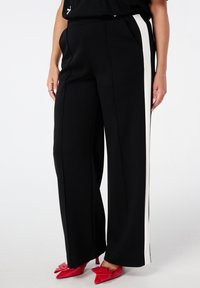 Femme portant un pantalon noir à large jambes avec des rayures blanches sur les côtés et des chaussures à talons aiguilles rouges avec un détail en nœud, se tenant devant un fond blanc.
