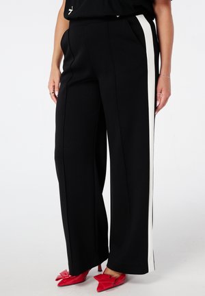 Vrouw in zwarte wijde broek met witte zijstrepen en rode puntige hakken met strikdetails, staand tegen een witte achtergrond.