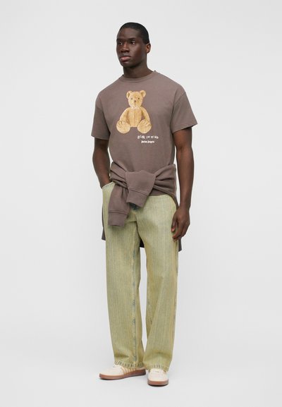Καφέ γραφικό t-shirt με σχέδιο αρκούδας, συνδυασμένο με ανοιχτό πράσινο, υφασμάτινο παντελόνι φαρδιάς γραμμής. Λευκά αθλητικά ολοκληρώνουν το σύνολο.